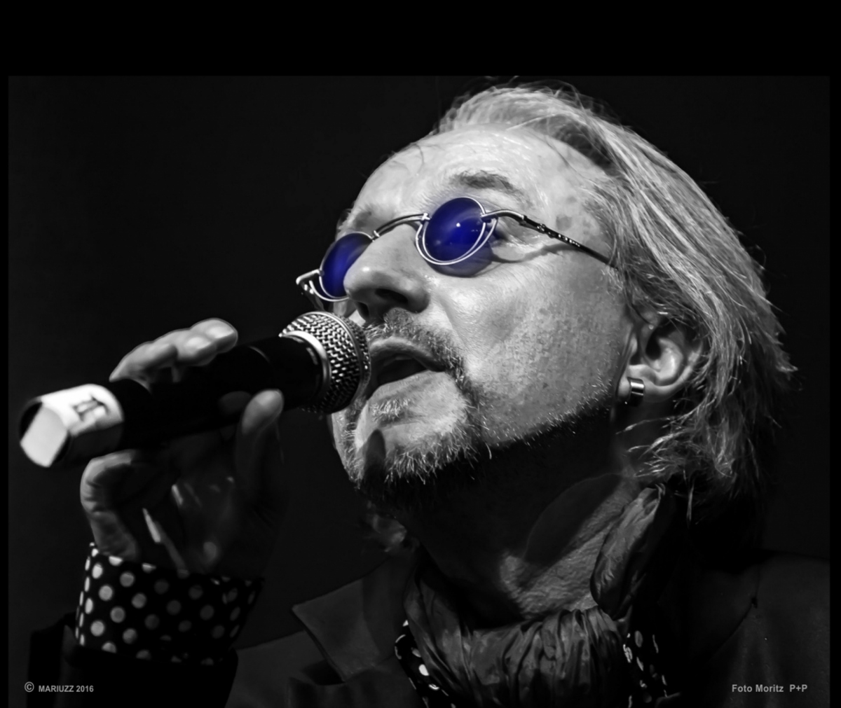 Das Gesicht eines Mannes mit Sonnebrille, kleine runde Gläser, singt in eine Mikrofon.