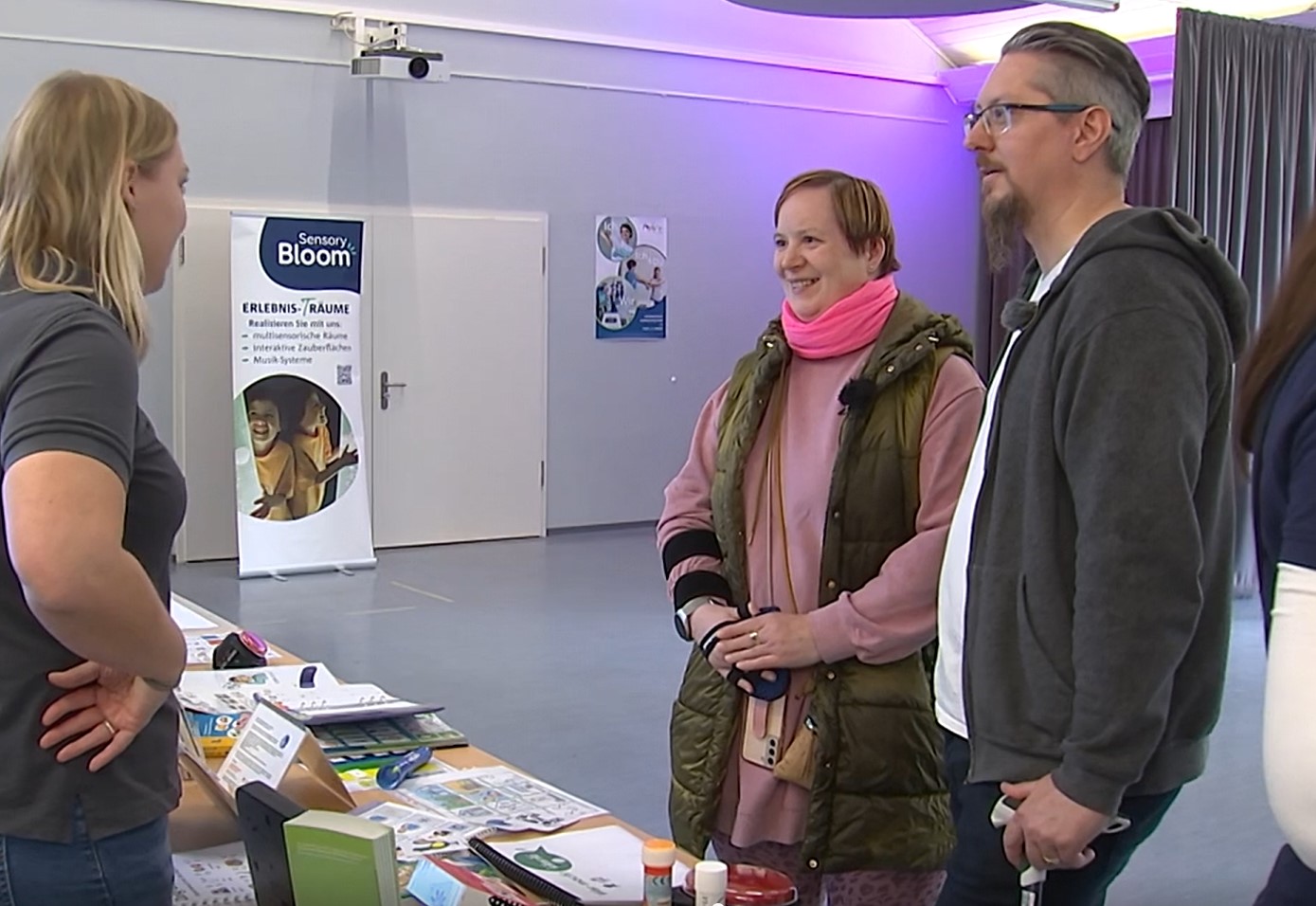 Eine Frau und ein Mann stehen in einem Saal an einem Tisch. Sie reden mit einer Frau. Auf dem Tisch liegen Flyer und Broschüren.
