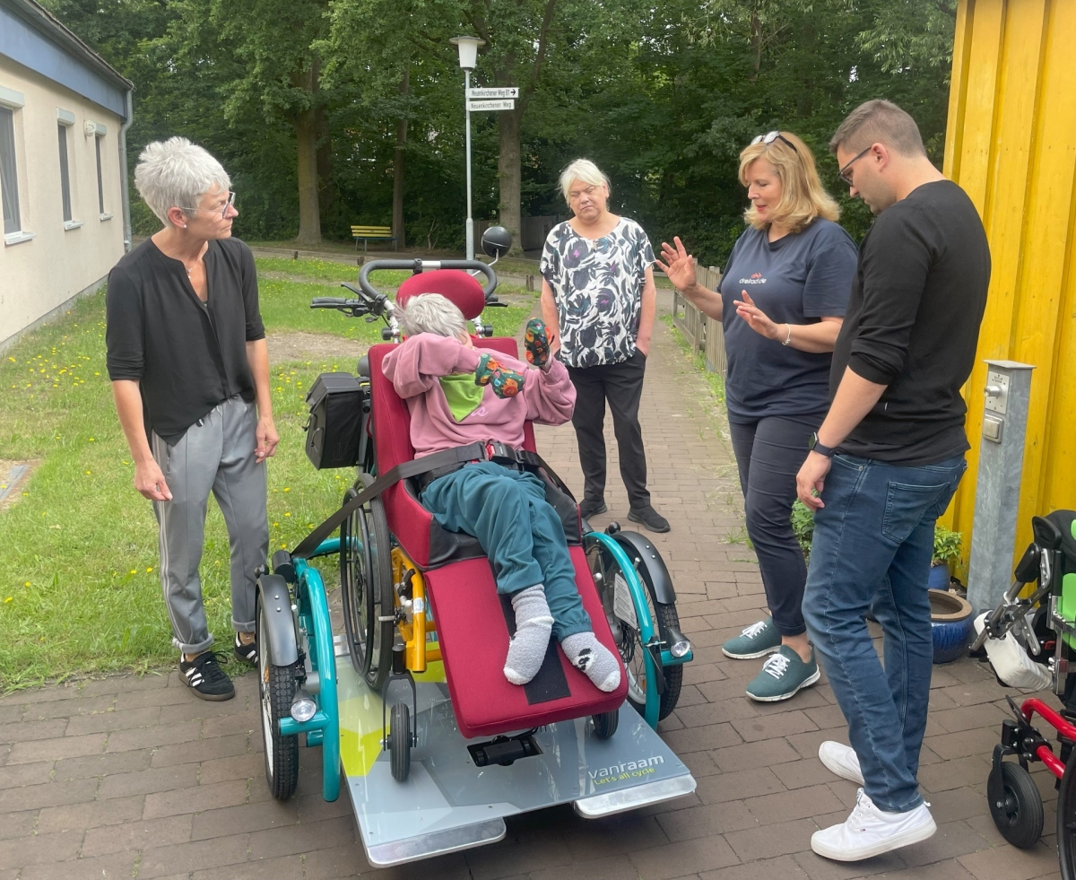 Mehrere Menschen schauen auf ein Fahrrad, das einen Rollstuhl mit einer Person transportiert. 