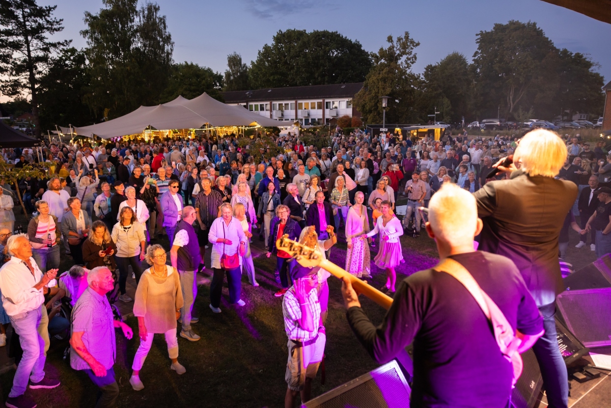 Hunderte Menschen stehen vor eine Bühne in der Dämmerung und tanzen. Musiker stehen auf der Bühne. 